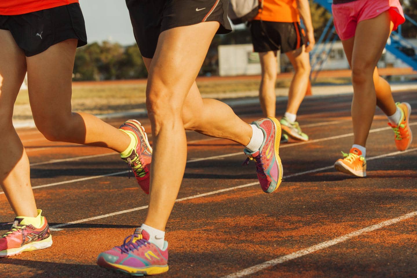 En gruppe løbere ses fra knæene og ned, mens de løber på en rød atletikbane. Benene er i bevægelse, og nogle bærer løbesko i forskellige farver.